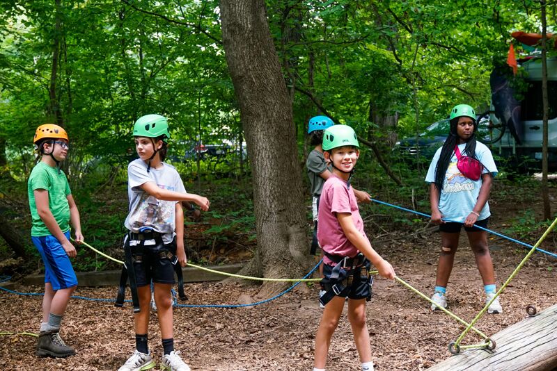 A group of children wearing helmets are participating in a ropes course activity in a forest. They are connected to ropes and harnesses, suggesting they are navigating an elevated obstacle course. The setting appears to be outdoors, with trees providing a natural backdrop for the adventurous activity.